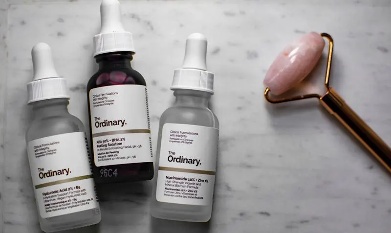 더오디너리 브랜드의 세럼 3종이 대리석 배경 위에 놓여 있으며, 나이아신아마이드와 히알루론산 제품이 함께 진열되어 있다. 옆에는 로즈 쿼츠 페이스 롤러가 배치되어 스킨케어 루틴의 감각적인 이미지를 완성한다. (A collection of The Ordinary skincare serums including niacinamide and hyaluronic acid on a marble surface, accompanied by a rose quartz face roller for an elegant skincare routine aesthetic.)