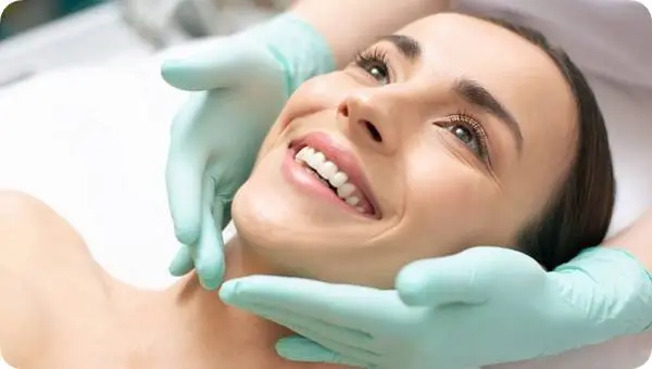 써마지 시술을 마친 여성이 편안하게 미소 짓는 모습. 의료진이 라텍스 장갑을 착용한 손으로 여성의 턱과 얼굴선을 부드럽게 잡아 피부 상태를 확인하고 있으며, 환자는 밝고 건강한 표정으로 만족스러워 보인다. (A smiling woman after Thermage treatment as a clinician wearing latex gloves gently examines her jawline and facial contours, showing satisfaction and healthy skin glow.)